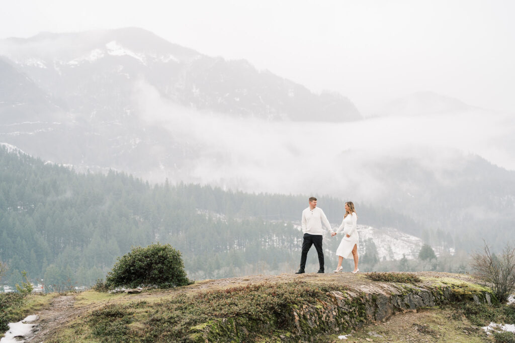 couple walking across a rocky outcropping with snowy winter mountains behind them during winter maternity photos near portland oregon