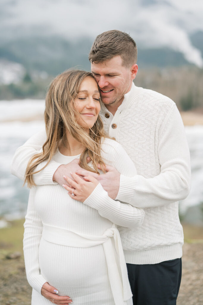 couple poses for PNW pregnancy picture outdoors in front of a snowy mountain and river